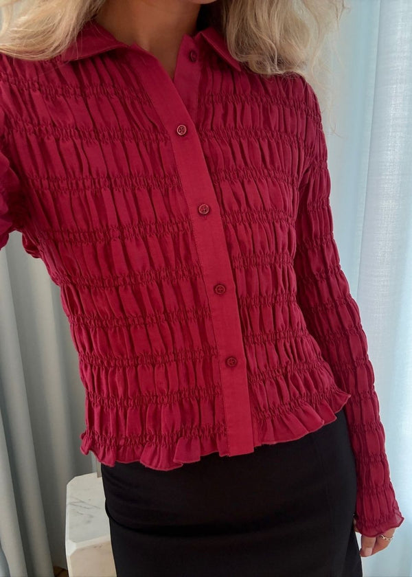 Langærmet tætsiddende top i mørkerød med smock. Posiemd shirt har en knappelukning fortil og skjorte krave. Modellen er 175 cm og har en str S/36 på.