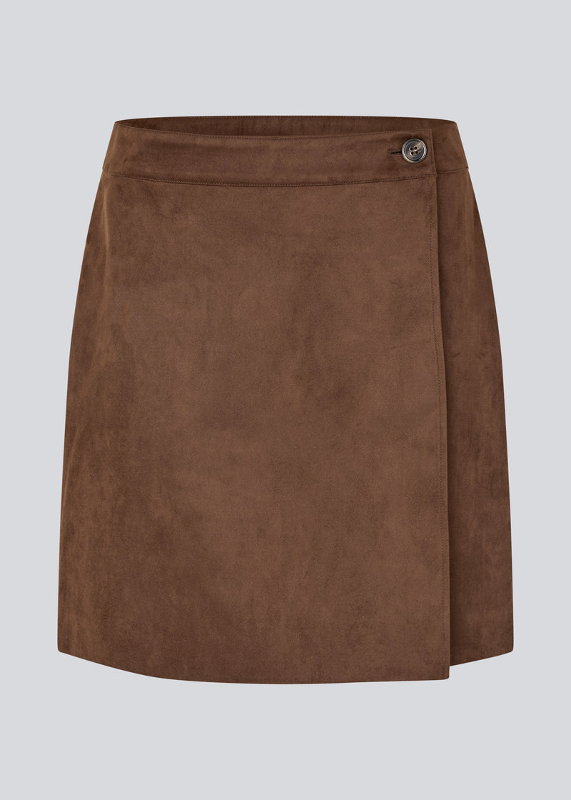 Kort brun nederdel i ruskindslook med elegant slåom design. Nederdelen lukkes med en dekorativ knap i siden, som tilføjer et stilrent og feminint touch. Det bløde materiale giver en behagelig pasform, og det enkle snit gør den nem at style til både hverdag og mere formelle lejligheder.