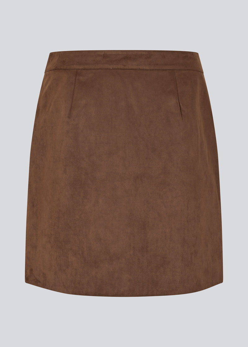 Kort brun nederdel i ruskindslook med elegant slåom design. Nederdelen lukkes med en dekorativ knap i siden, som tilføjer et stilrent og feminint touch. Det bløde materiale giver en behagelig pasform, og det enkle snit gør den nem at style til både hverdag og mere formelle lejligheder.