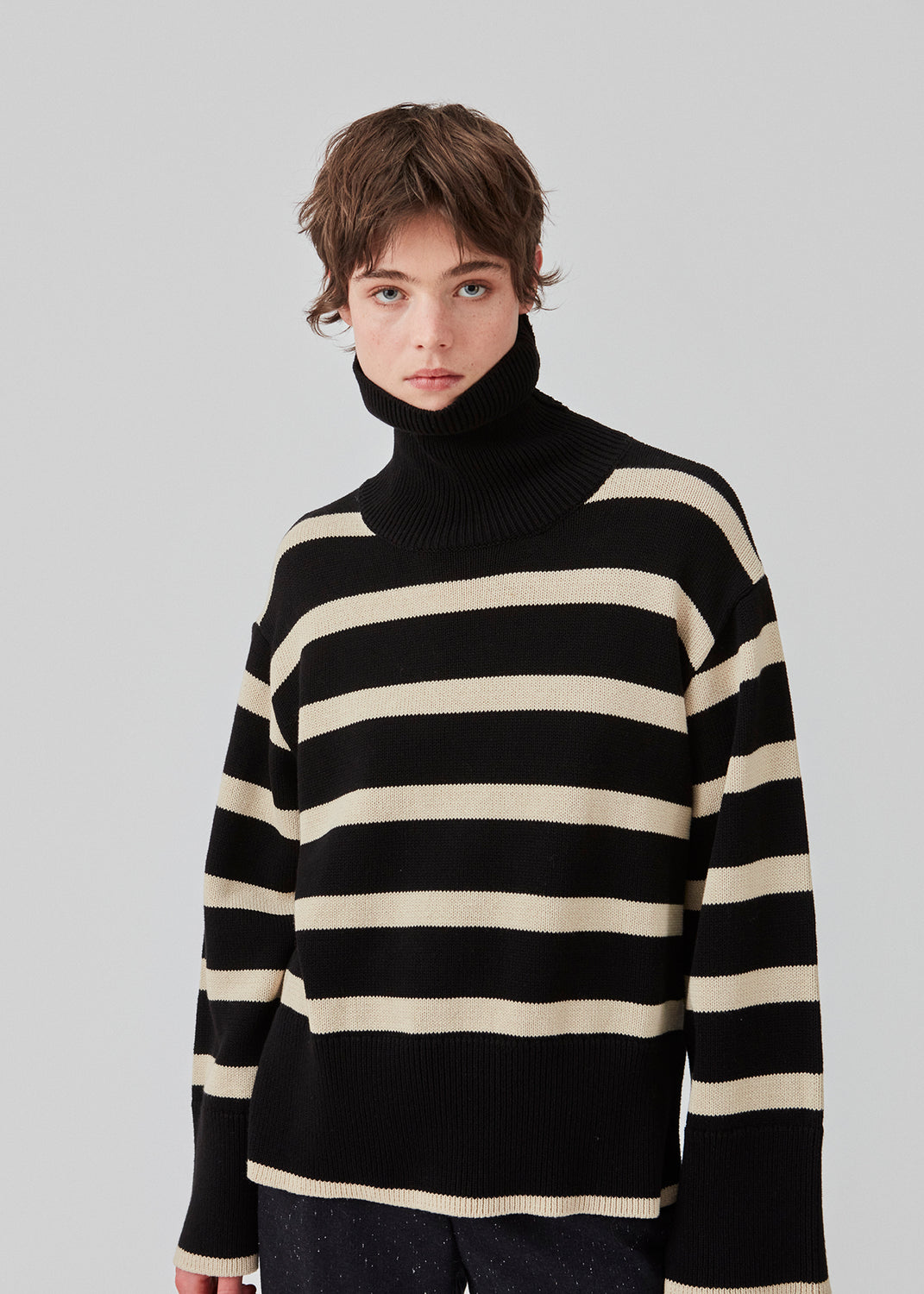Finstrikket, oversized trøje i bomuldsstrik i sort med beige striber. CorbinMD stripe t-neck har en ribstrikket rullekrave, lange brede ærmer og brede ribkanter ved ærmer og forneden. Modellen er 177 cm og har en størrelse S/36 på.