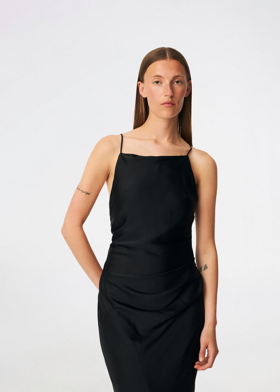 DanillaMD Dress er en lang satin-kjole med en elegant, figurnær silhuet. Designet har en lige halsudskæring, tynde stropper og draperede detaljer i siden, som skaber et blødt fald. Den åbne ryg med fine krydsende stropper tilfører et feminint udtryk. Style den med hæle og enkle accessories til særlige anledninger. Modellen er 175 cm høj og bruger størrelse S/36.