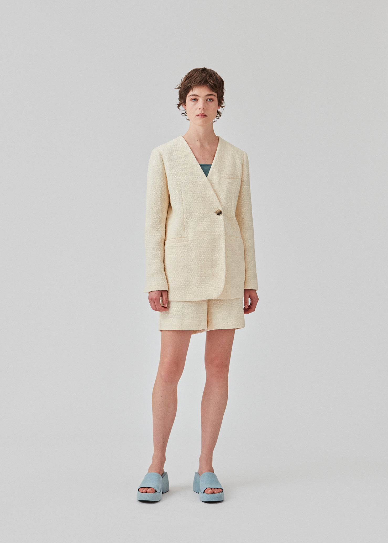 DimeMD blazer er designet i et tekstureret bomuldsmix med foer. Blazeren er uden krave og revers, men har en asymmetrisk wrap-detalje med en enkelt synlig knap. Modellen er 177 cm og har en størrelse S/36 på.  Style blazeren med matchende shorts: DimeMD shorts.