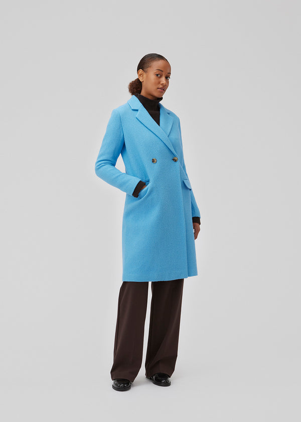 Dobbeltradet frakke med revers og knapper foran. EdinMD coat er mellemlang med lange ærmer, forlommer med flap og slids bagpå. Med foer. Modellen er 175 cm og har en størrelse S/36 på.