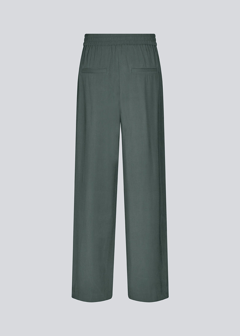 FanyaMD pants i mørkegrøn har et herre-inspireret look med lige, brede ben, høj talje med lynlåsgylp og knap og elastik bagpå. Dobbeltlæg foran og sidelommer. Modellen er 175 cm og har en størrelse S/36 på.