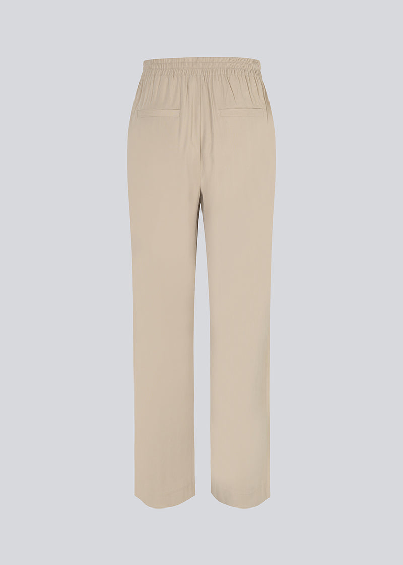 FanyaMD pants i lys beige har et herre-inspireret look med lige, brede ben, høj talje med lynlåsgylp og knap og elastik bagpå. Dobbeltlæg foran og sidelommer. Modellen er 175 cm og har en størrelse S/36 på.