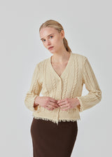 Beige cardigan i kabelstrik med bomuld. Oversized og let cropped pasform med lange ærmer og rå kanter. FinaMD cardigan har v-udskæring og knapper foran. Modellen er 175 cm og har en størrelse S/36 på.