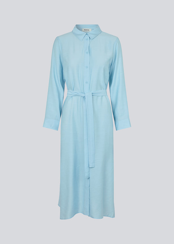 Midi-kjole i lyseblå i vævet kvalitet med krave, knapper foran, aftageligt bindebælte i taljen og slidser i siden. Lange ærmer med manchet. Modellen er 175 cm og har en størrelse S/36 på.