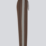 Løse brune bukser med bred benvidde og afslappet pasform. Bukserne har elastisk linning med snøre, der sikrer en behagelig og justerbar talje. Langs siderne løber hvide striber, som tilføjer et sporty og moderne twist til det ellers klassiske design. Perfekte til både afslappet hverdagsbrug og mere stilfulde outfits – nemme at style med alt fra t-shirts til skjorter.
