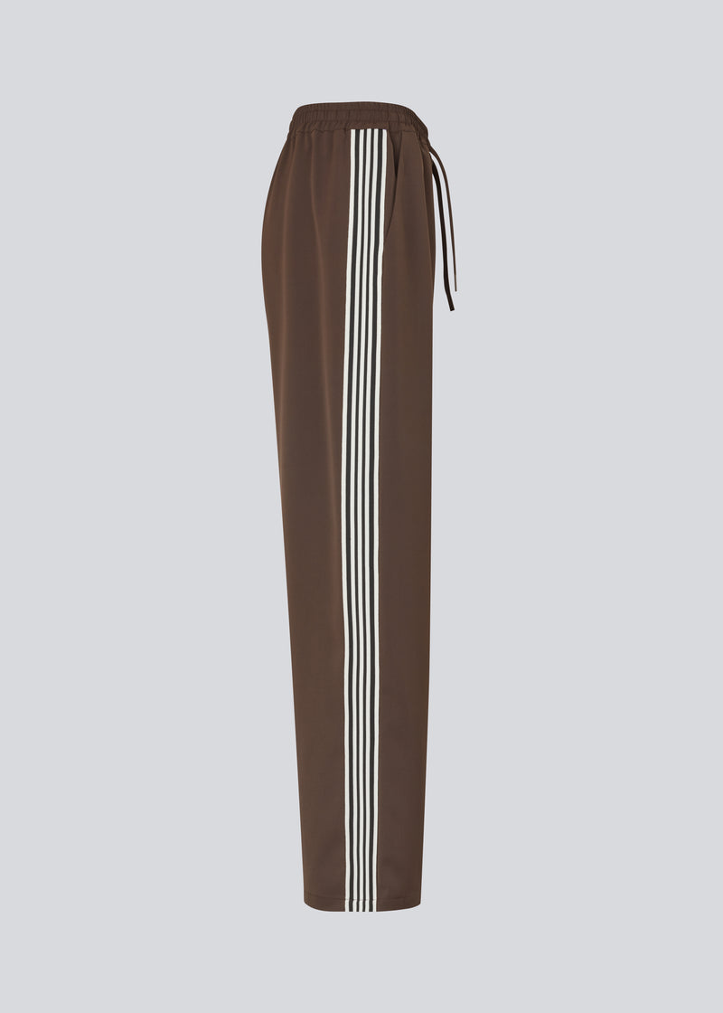 Løse brune bukser med bred benvidde og afslappet pasform. Bukserne har elastisk linning med snøre, der sikrer en behagelig og justerbar talje. Langs siderne løber hvide striber, som tilføjer et sporty og moderne twist til det ellers klassiske design. Perfekte til både afslappet hverdagsbrug og mere stilfulde outfits – nemme at style med alt fra t-shirts til skjorter.
