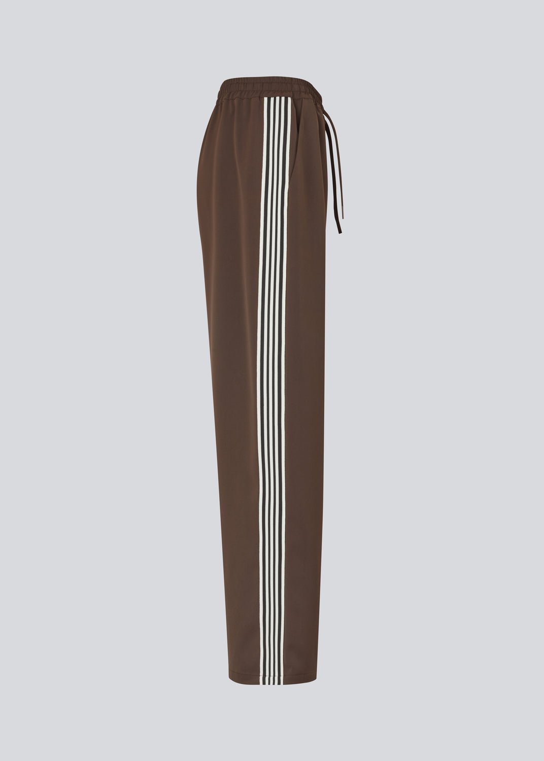Løse brune bukser med bred benvidde og afslappet pasform. Bukserne har elastisk linning med snøre, der sikrer en behagelig og justerbar talje. Langs siderne løber hvide striber, som tilføjer et sporty og moderne twist til det ellers klassiske design. Perfekte til både afslappet hverdagsbrug og mere stilfulde outfits – nemme at style med alt fra t-shirts til skjorter.