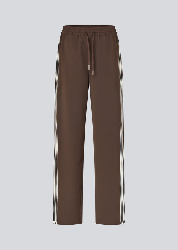 Løse brune bukser med bred benvidde og afslappet pasform. Bukserne har elastisk linning med snøre, der sikrer en behagelig og justerbar talje. Langs siderne løber hvide striber, som tilføjer et sporty og moderne twist til det ellers klassiske design. Perfekte til både afslappet hverdagsbrug og mere stilfulde outfits – nemme at style med alt fra t-shirts til skjorter.