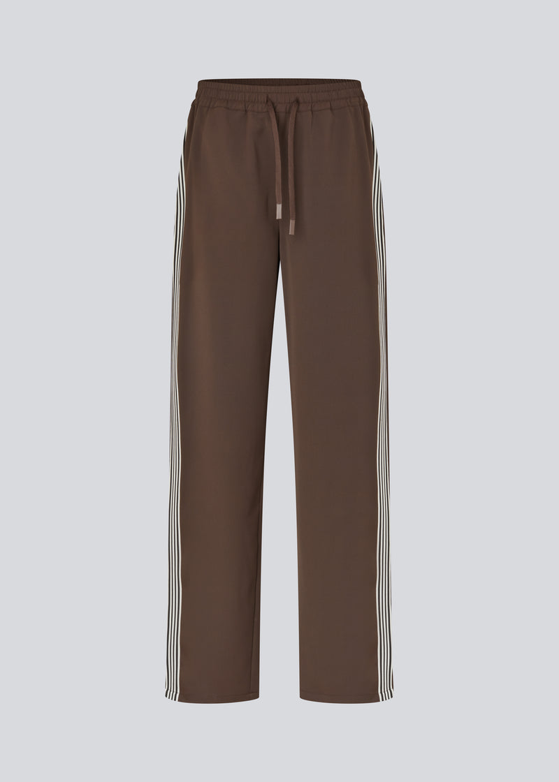 Løse brune bukser med bred benvidde og afslappet pasform. Bukserne har elastisk linning med snøre, der sikrer en behagelig og justerbar talje. Langs siderne løber hvide striber, som tilføjer et sporty og moderne twist til det ellers klassiske design. Perfekte til både afslappet hverdagsbrug og mere stilfulde outfits – nemme at style med alt fra t-shirts til skjorter.