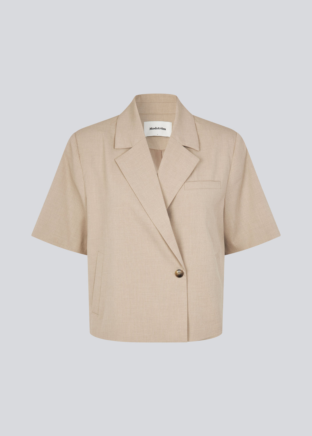 Kortærmet beige blazer med enkeltradet lukning. TuesdayMD SS Blazer har en krydsende knaplukning, krave, sidelommer og en falsk brystlomme – et moderne twist på en tidløs klassiker. Kan bæres lukket som en elegant top eller åben over en let bluse for et mere afslappet udtryk.&nbsp;