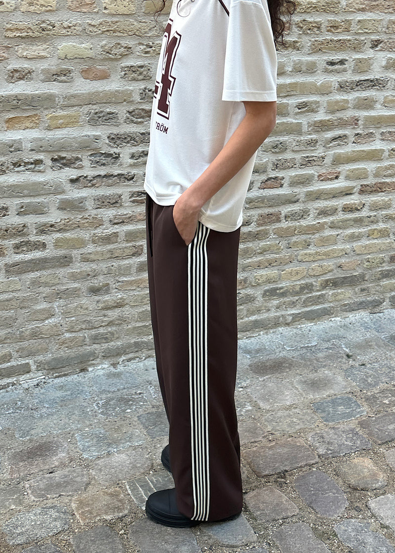 Løse brune bukser med bred benvidde og afslappet pasform. Bukserne har elastisk linning med snøre, der sikrer en behagelig og justerbar talje. Langs siderne løber hvide striber, som tilføjer et sporty og moderne twist til det ellers klassiske design. Perfekte til både afslappet hverdagsbrug og mere stilfulde outfits – nemme at style med alt fra t-shirts til skjorter.