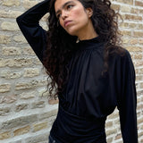 Elegant sort bluse med høj hals og lange ærmer. Blusen er designet med fine rynkedetaljer ved brystet og taljen, som skaber en flatterende silhuet. Den har en diskret knaplukning bagpå i nakken, som giver et sofistikeret og feminint udtryk. Det strækbare stof sikrer en behagelig pasform, mens det draperede snit tilføjer ekstra elegance. Perfekt til både hverdagsbrug og festlige lejligheder.