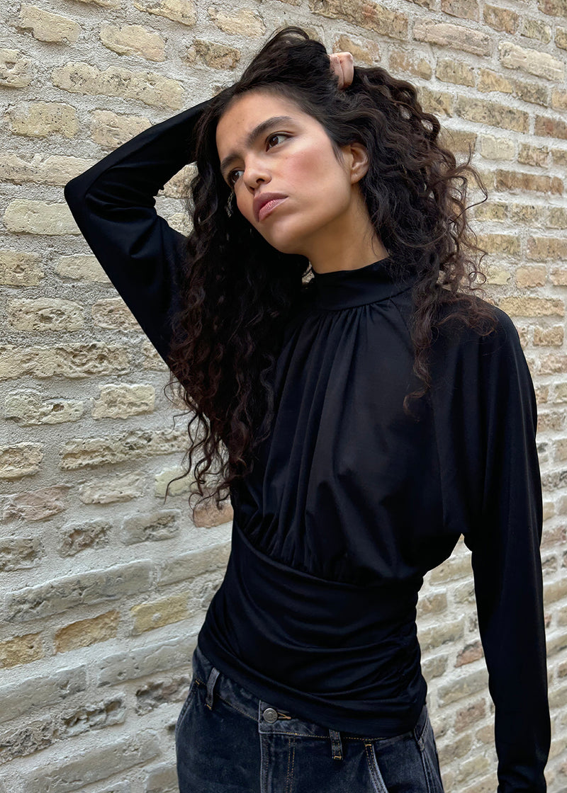 Elegant sort bluse med høj hals og lange ærmer. Blusen er designet med fine rynkedetaljer ved brystet og taljen, som skaber en flatterende silhuet. Den har en diskret knaplukning bagpå i nakken, som giver et sofistikeret og feminint udtryk. Det strækbare stof sikrer en behagelig pasform, mens det draperede snit tilføjer ekstra elegance. Perfekt til både hverdagsbrug og festlige lejligheder.