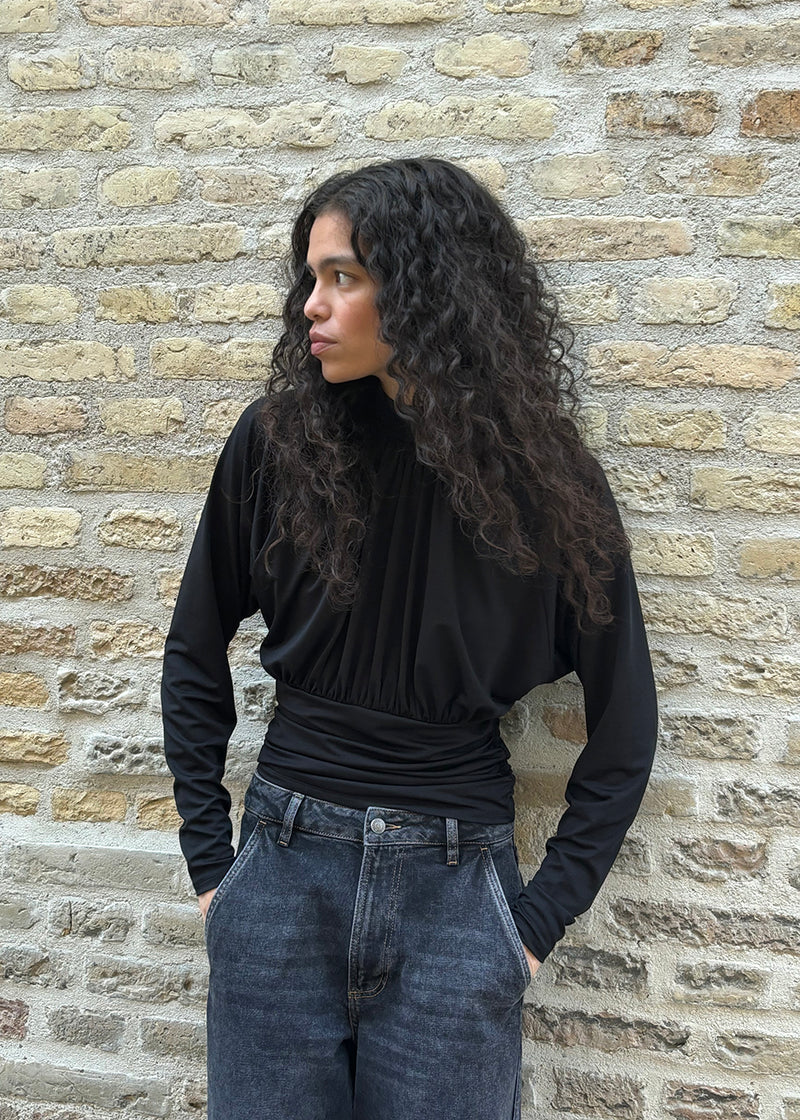 Elegant sort bluse med høj hals og lange ærmer. Blusen er designet med fine rynkedetaljer ved brystet og taljen, som skaber en flatterende silhuet. Den har en diskret knaplukning bagpå i nakken, som giver et sofistikeret og feminint udtryk. Det strækbare stof sikrer en behagelig pasform, mens det draperede snit tilføjer ekstra elegance. Perfekt til både hverdagsbrug og festlige lejligheder.