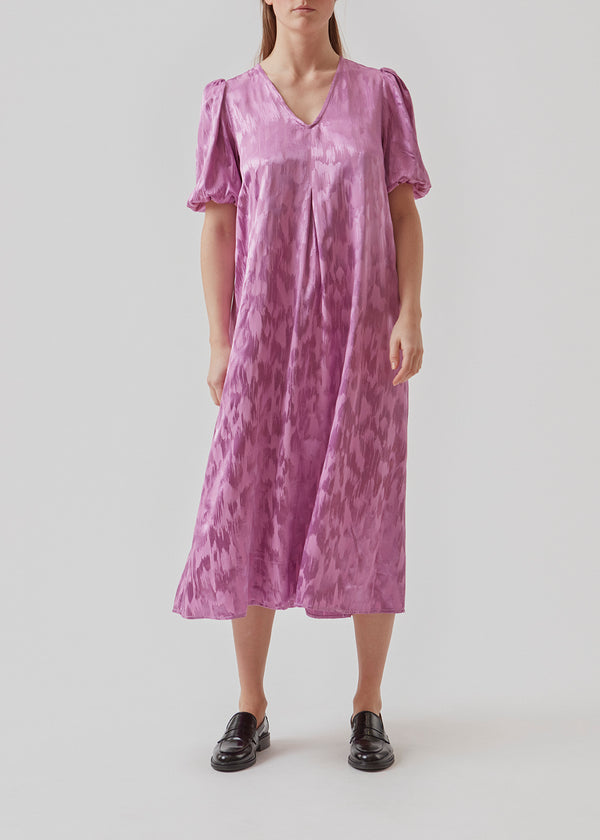 Mellemlang kjole med korte pufærmer med elastik. AbigaleMD dress har et skinnende mønster, v-udskæring i halsen og wienerlæg foran.