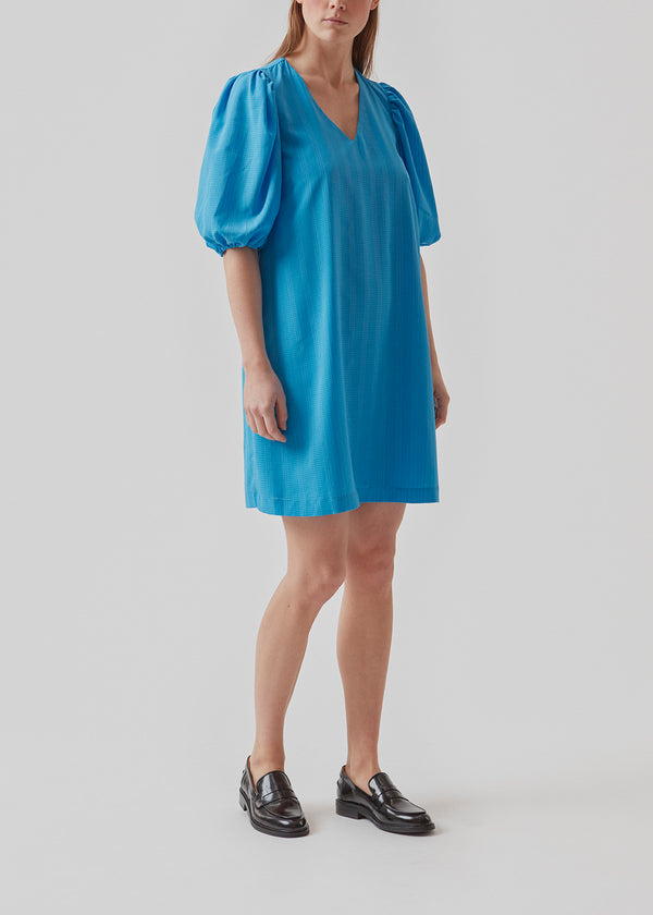 Kort kjole i blå med afslappet pasform og v-udskæring. Ærmerne på AshaMD dress er korte og voluminøse med elastikkant.  Materiale: 100% Polyester  Foer: 100% Polyester