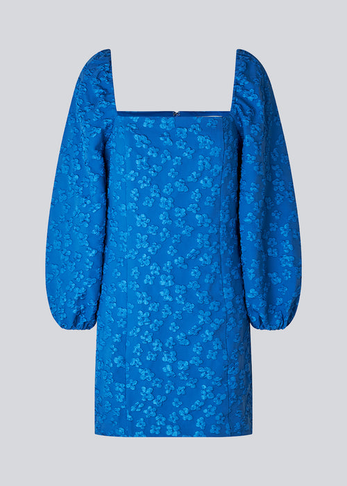 Kort kjole i blå med all-over blomstermotiv. AtiraMD dress har lange ballonærmer med en mere kropsnær krop. Halsudskæringen er firkantet både foran og bagpå.  Modellen er 177 cm og har en størrelse S/36 på.