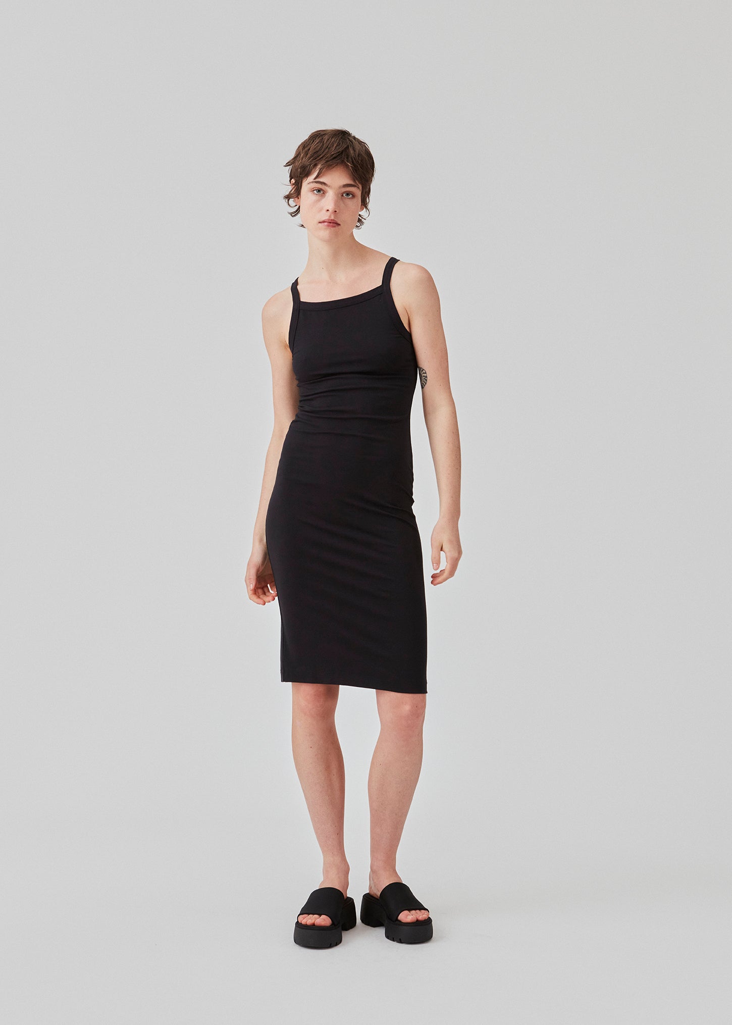 Tætsiddende basic sort kjole i en økologisk bomuldsjersey. DaeMD dress er uden ærmer og har en høj og lige halsudskæring. Kjolen skærer under knæene. Modellen er 177 cm og har en størrelse S/36 på.