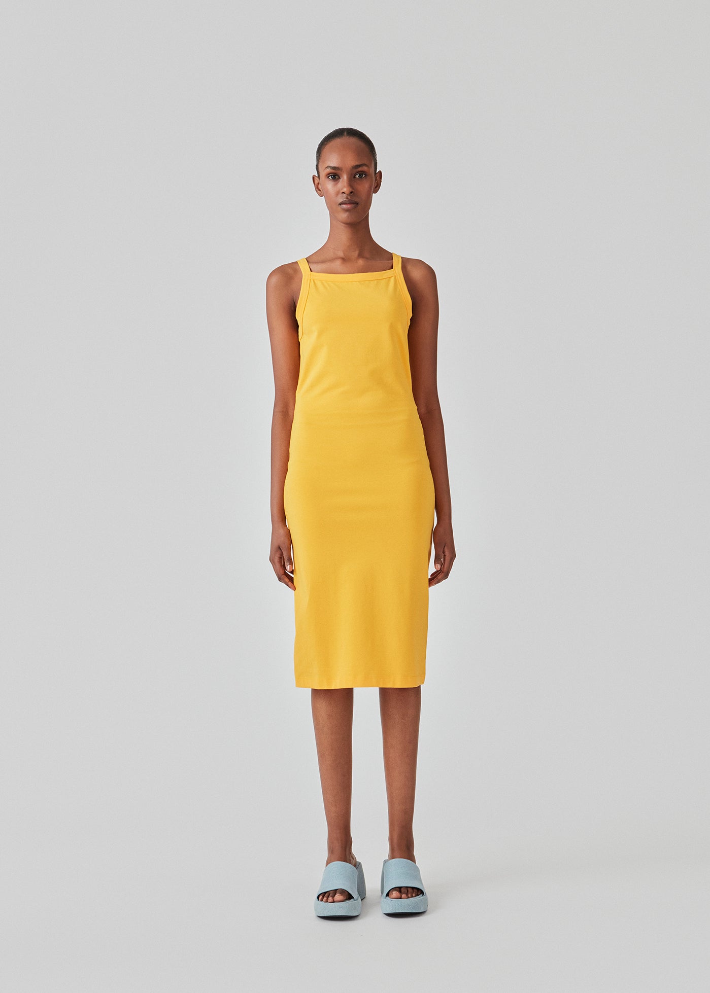 Tætsiddende basic gul kjole i en økologisk bomuldsjersey. DaeMD dress er uden ærmer og har en høj og lige halsudskæring. Kjolen skærer under knæene. Modellen er 177 cm og har en størrelse S/36 på.