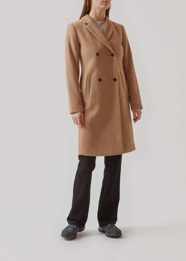 Smuk knælang uldfrakke i lys brun. Odelia coat bliver lukket fortil af fire knapper og er taljeret, hvilket giver et feminint udtryk. På grund af den høje kvalitet af uld, vil den være det oplagte valg til både efterår og de milde vintre.