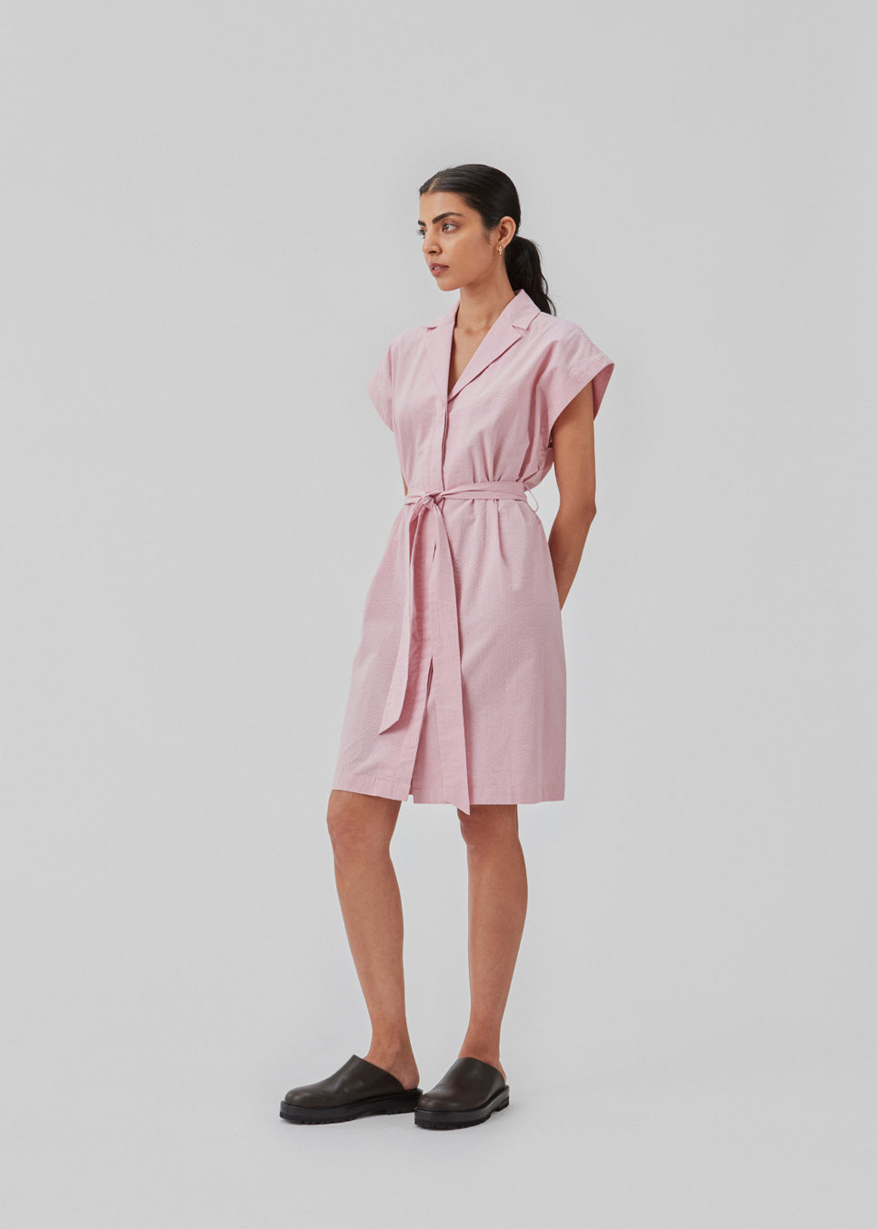Panne dress er en bomuldskjole i lyserød med smuk silhouet. Kjolen har desuden en fin krave, bindebånd og et ærmeløst design med vide, firkantede skuldre. Modellen er 173 cm og har en størrelse S/36 på.