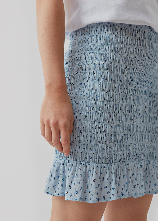 Porscha print skirt er en kort nederdel med elastisk smock over hele skørtet, som afsluttes med en bred flæsekant. Det blomsterprintede materiale er en EcoVero viskosekvalitet. Modellen er 173 cm og har en størrelse S/36 på